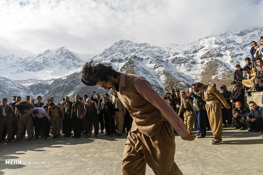 Bailarín con ropa kurda marrón se inclina ante un círculo de participantes y espectadores con montañas nevadas detrás