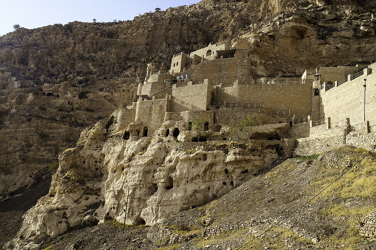 Stone terraces and buildings of Rabban Hormizd Monastery against a steep rock face, with cave openings below