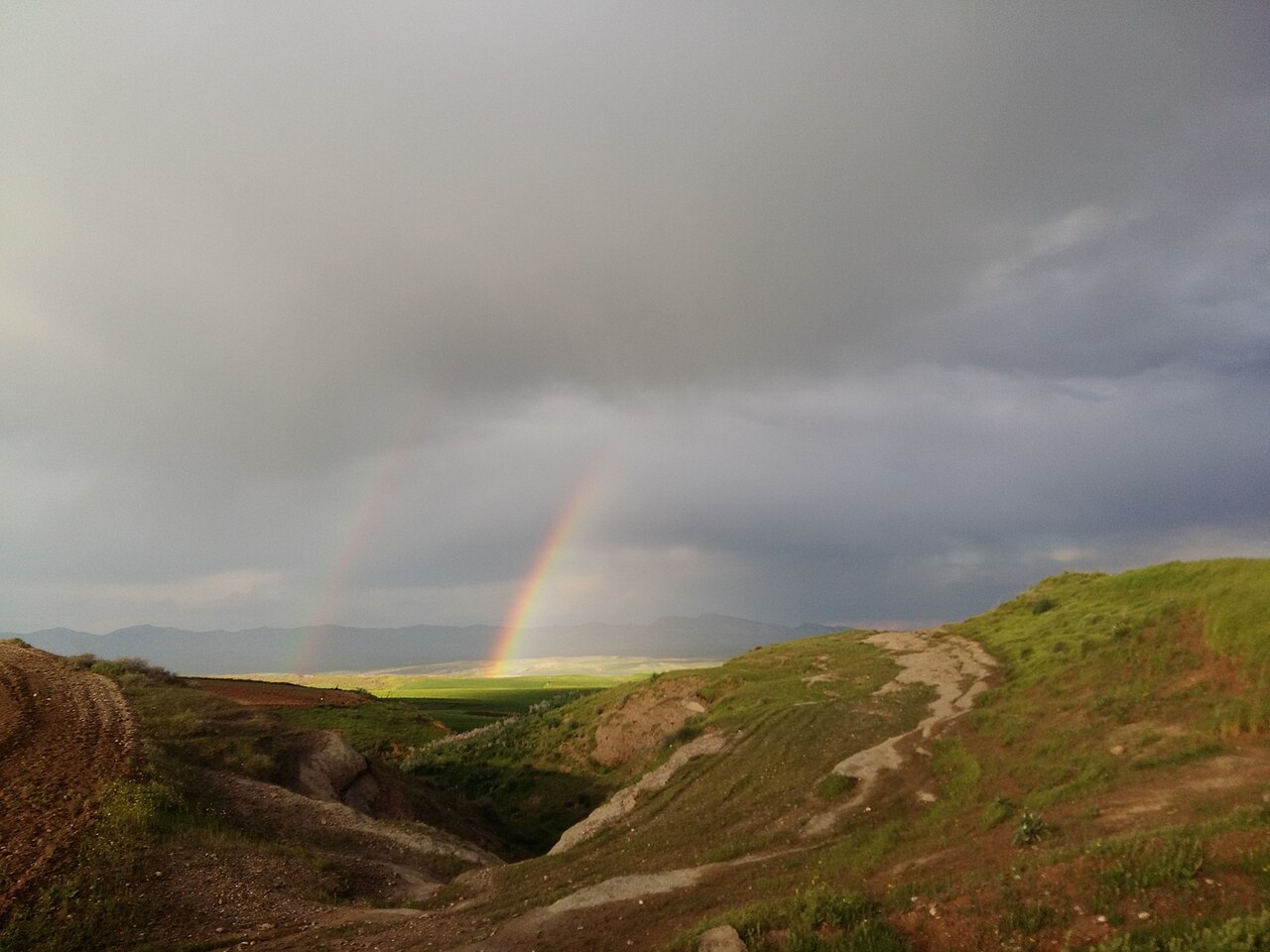 A bright rainbow and a fainter second arc beneath grey clouds above green fields and eroded hills at Banimaran near Koya