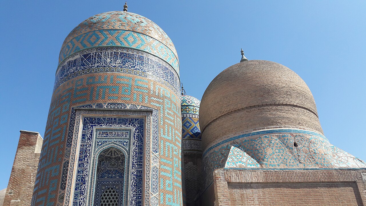 Cilindrische graftoren met turquoise patronen en aangrenzende bakstenen koepel in het Safi-heiligdom in Ardabil onder blauwe lucht