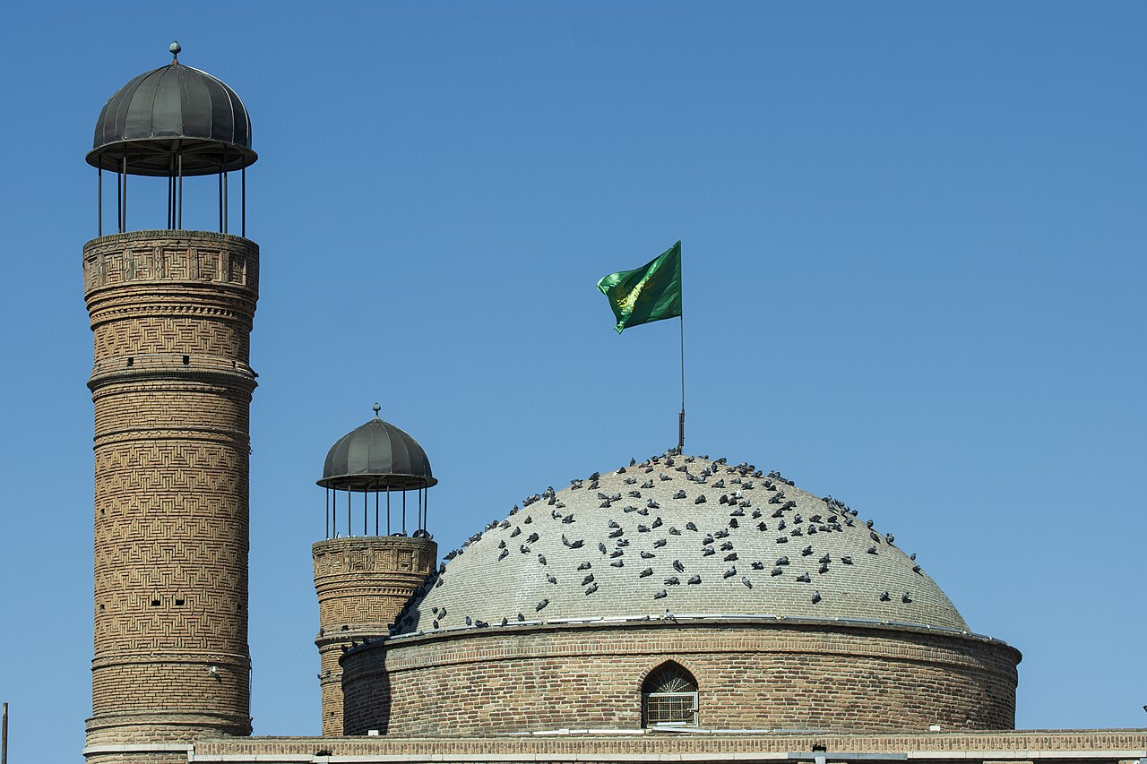 Saheb al-Amr Mosque in Tabriz with two patterned brick minarets, pigeons on a pale dome and a green flag against a clear blue sky