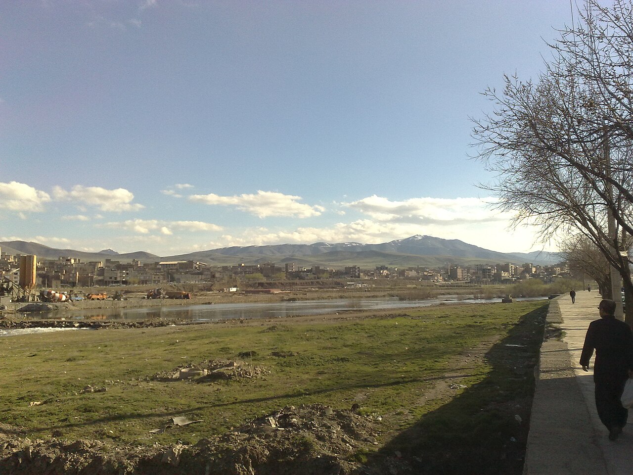 Saqqez seen across its river, with low buildings, mountains in the distance and a tree-lined footpath in the foreground