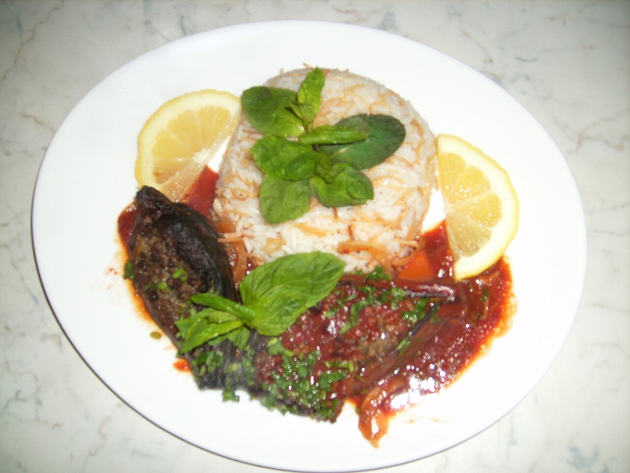 Met vlees gevulde aubergine in rode saus naast rijst met vermicelli, munt en citroenschijfjes op een wit bord