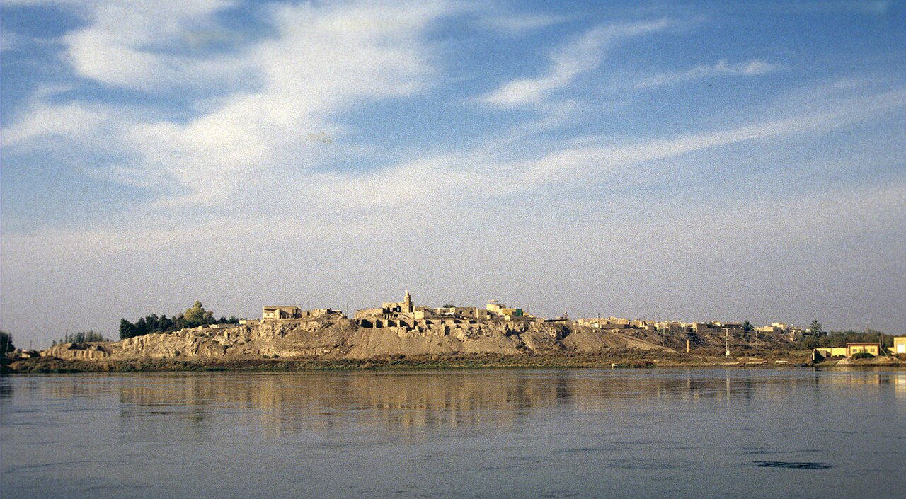 Archeologische heuvel van Terqa met gebouwen op de kam, gezien over een brede rivier onder wolken