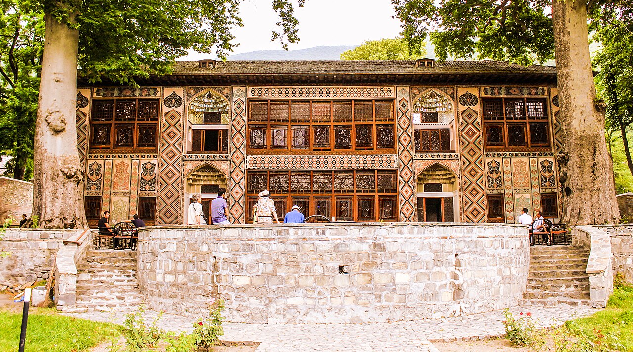Decorated two-storey façade of Sheki’s Khan Palace with wooden lattice windows, geometric painted panels, visitors and large trees in front