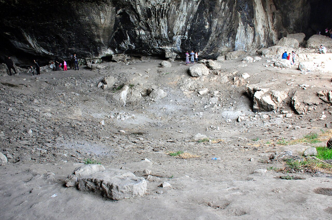 Visitors standing beneath Shanidar Cave’s high dark rock overhang, with large boulders and archaeological cuts across the dusty floor