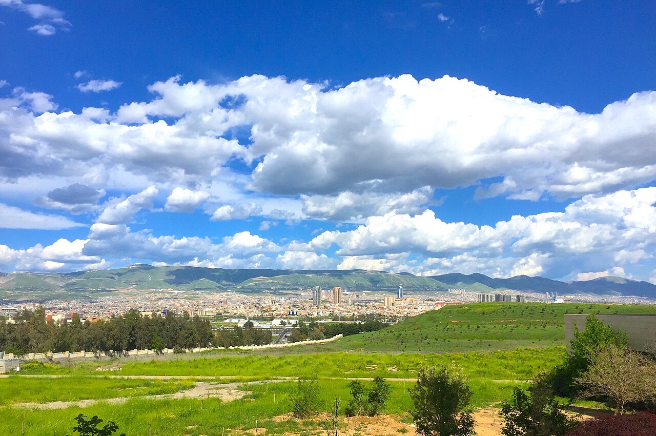 Modern Sulaimani spread across a valley beyond green fields, with mountains and large clouds behind the city