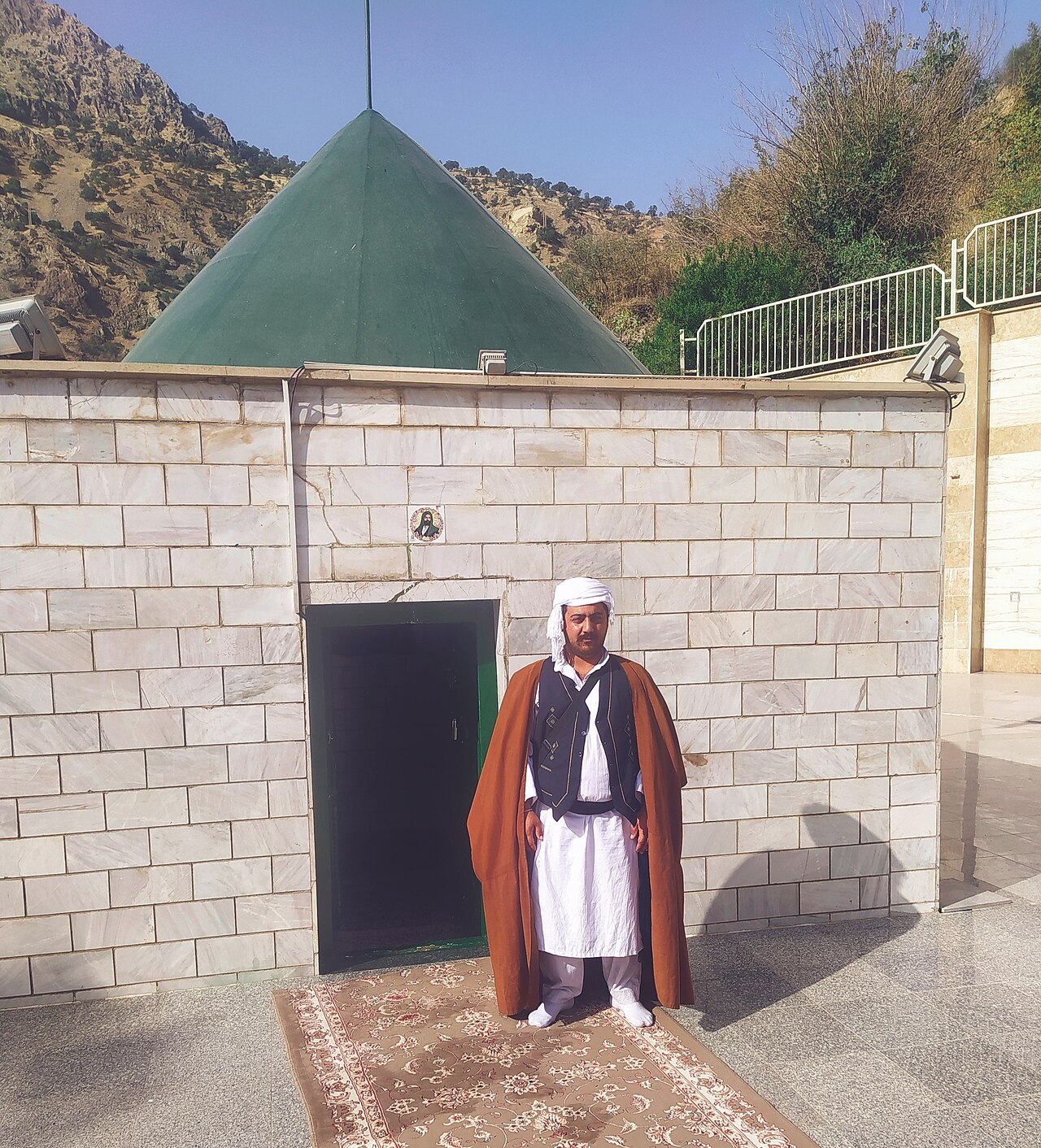 A visitor in a white headcloth and brown cloak outside the green-roofed shrine of Sultan Sahak