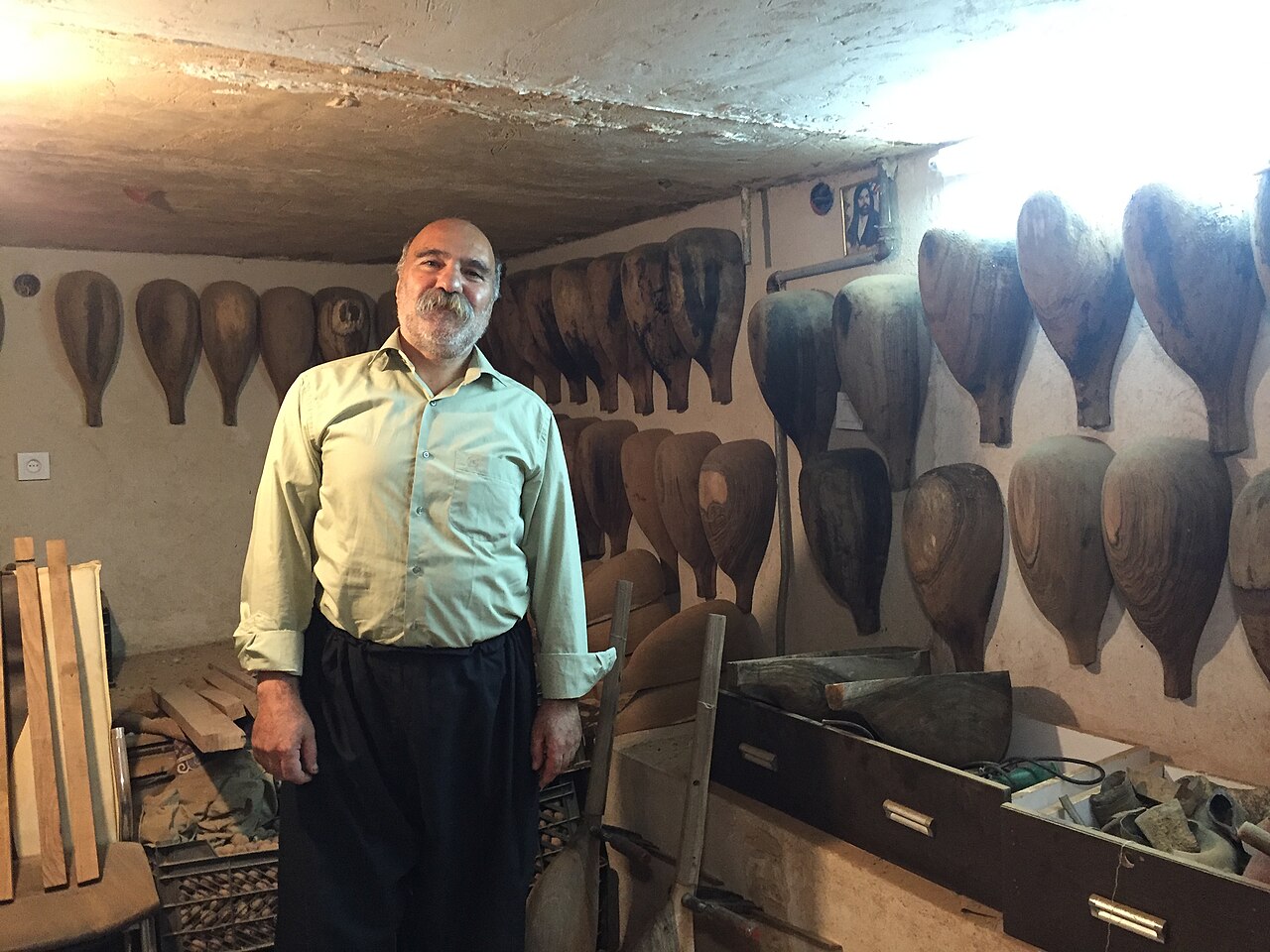 Tanbur maker in a pale green shirt standing in a workshop with unfinished pear-shaped wooden instrument bodies hanging on the walls and tools on a bench