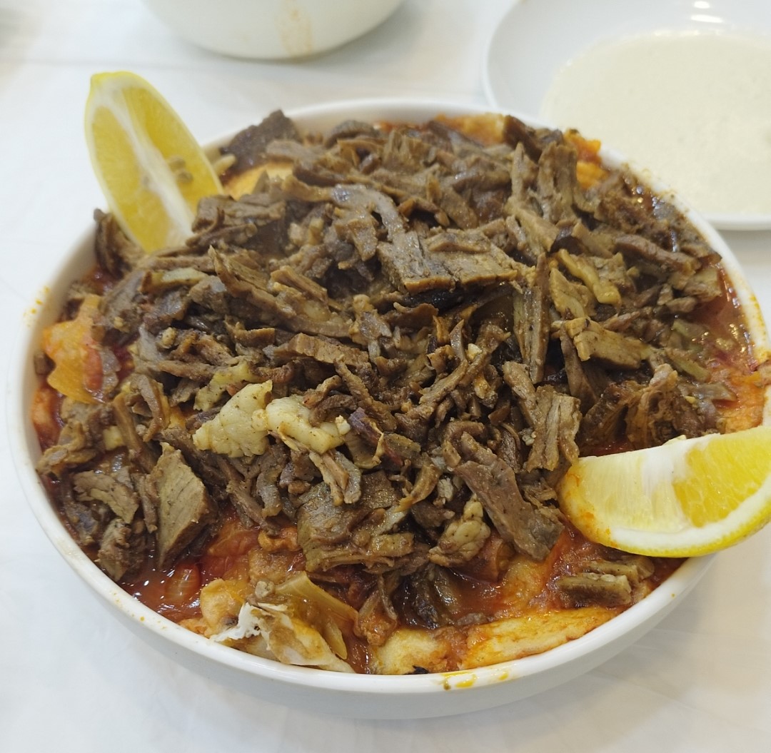 Sliced meat piled over bread and red sauce in a white bowl with two lemon wedges