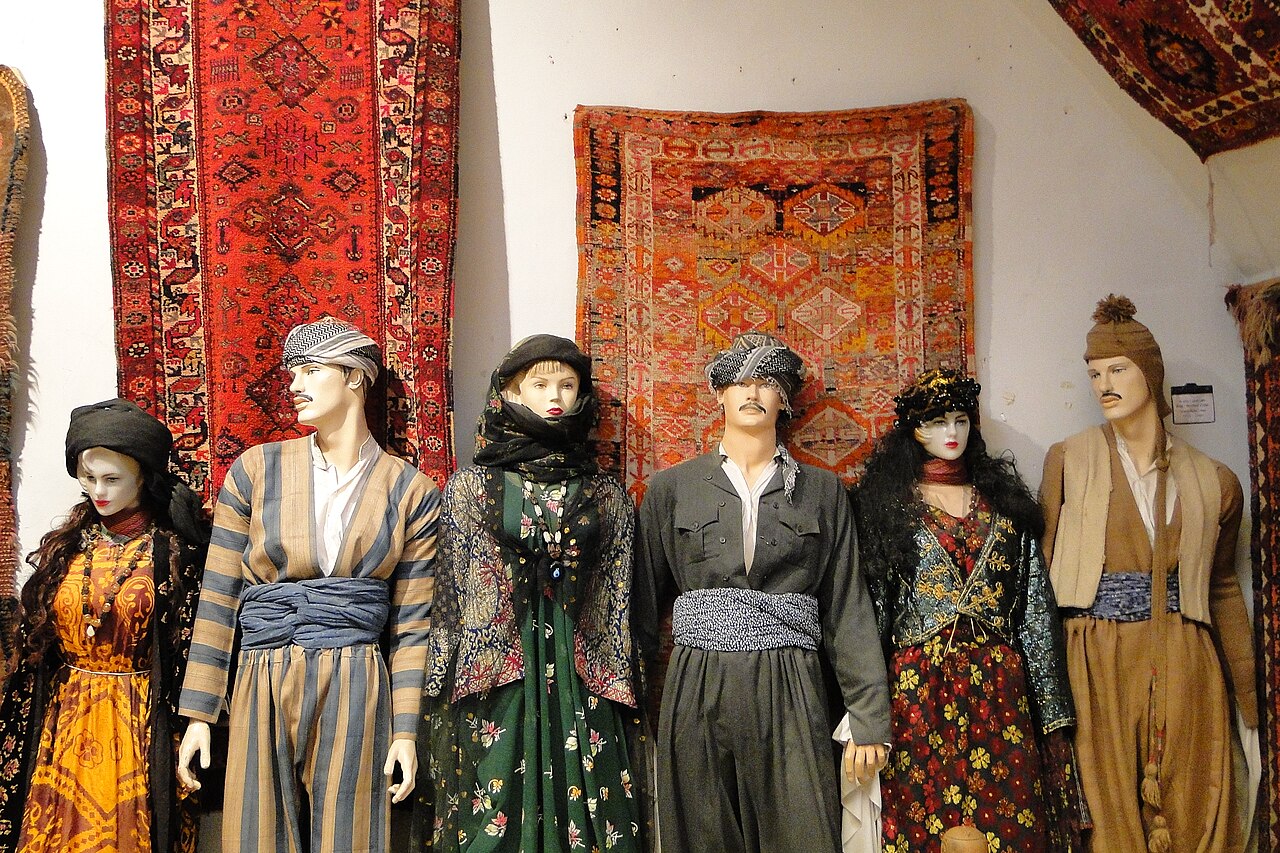 Six mannequins wearing varied Kurdish dresses, jackets, wide trousers, sashes and head coverings before patterned carpets in a museum