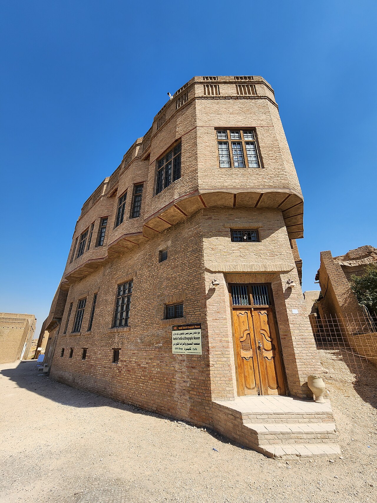 Tall brick facade of the Kurdish Textile Museum with projecting upper windows, a wooden double door, entrance steps and a museum sign