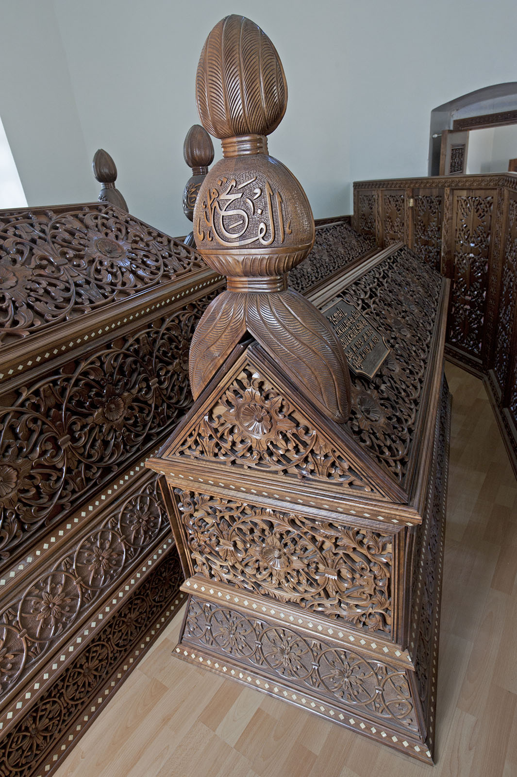Carved wooden cenotaphs inside the Tillo mausoleum, with floral latticework and a tall finial carrying Arabic calligraphy