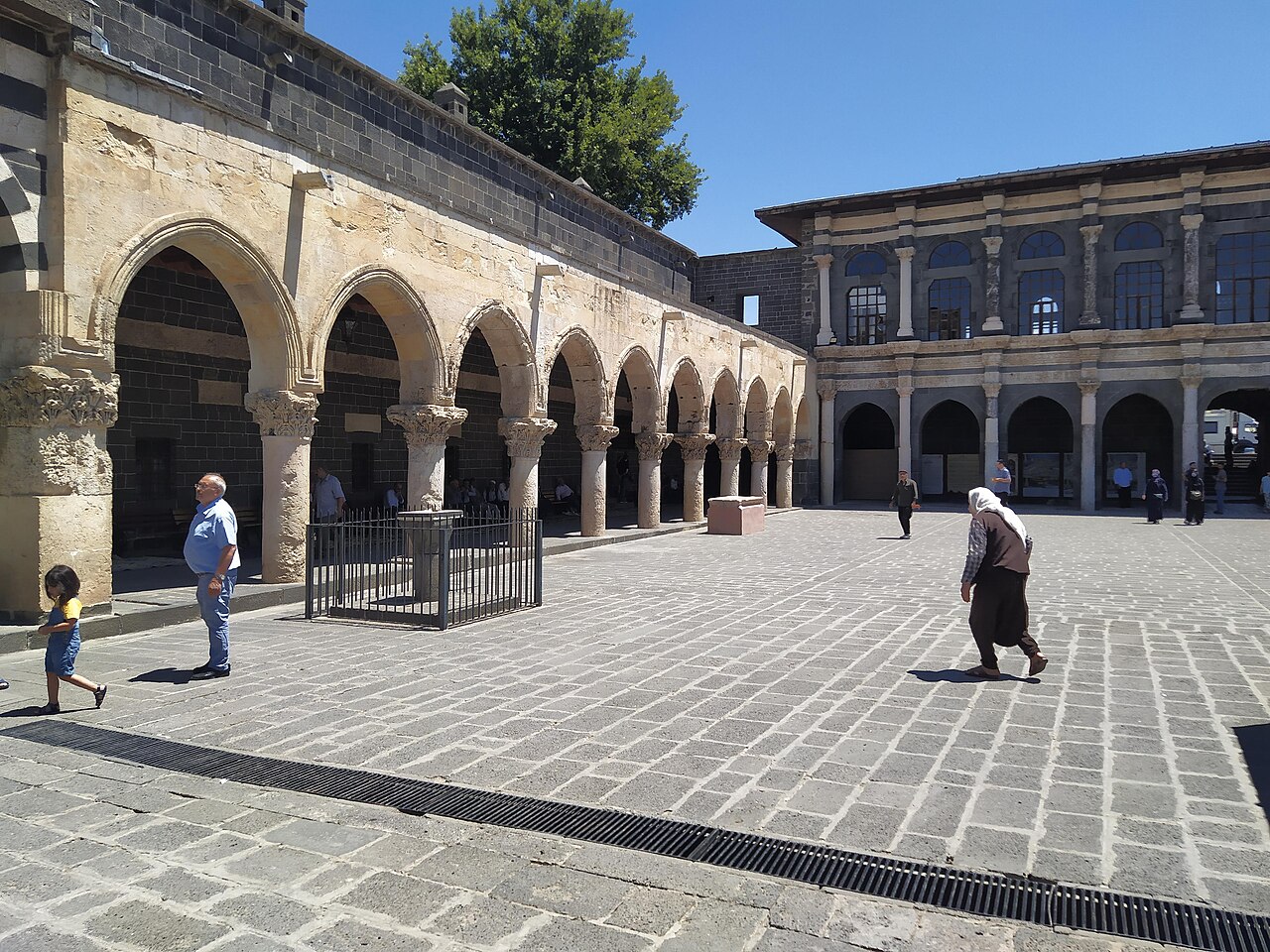Visitors crossing the paved Great Mosque courtyard beside a columned arcade with carved capitals and an adjoining two-storey façade