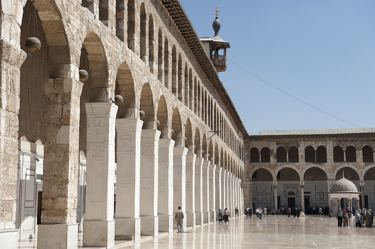 Visitors beside the stone arcades and reflective paving of the Umayyad Mosque courtyard in Damascus