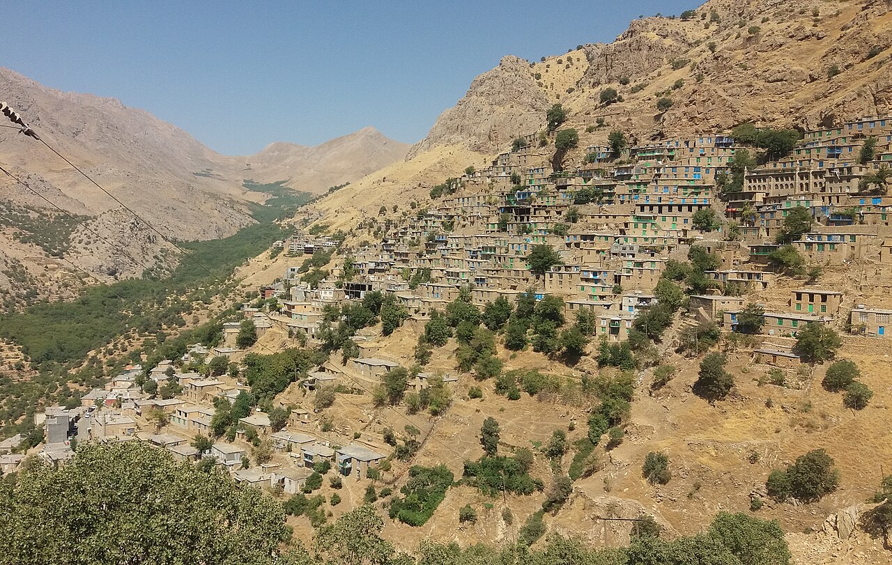 Edificios de piedra con ventanas azules y verdes escalonados en ladera seca sobre valle arbolado en Uraman Takht