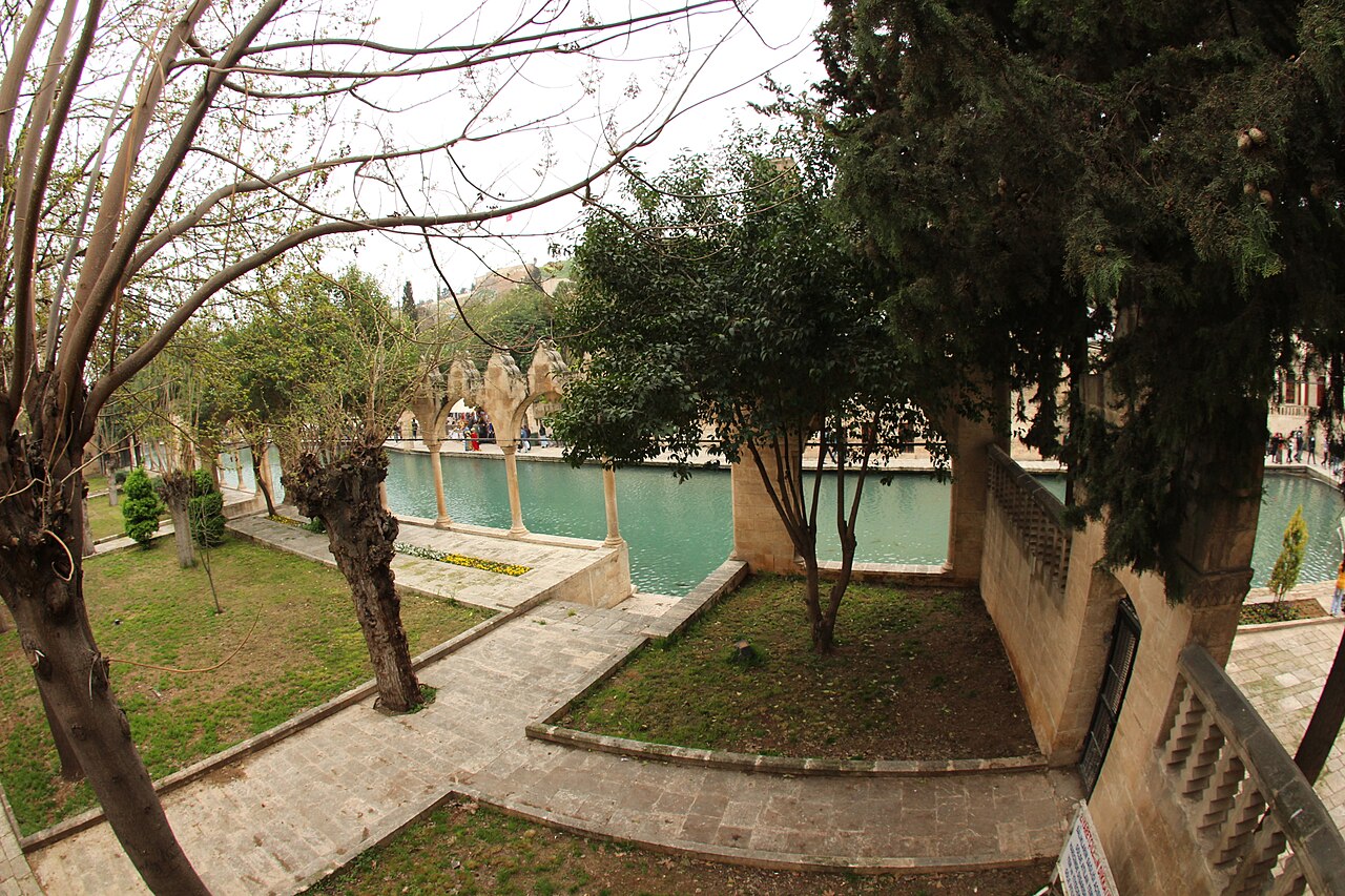 Trees and stone paths beside the turquoise Balıklıgöl pool in Urfa, with a small stone arcade at the water’s edge
