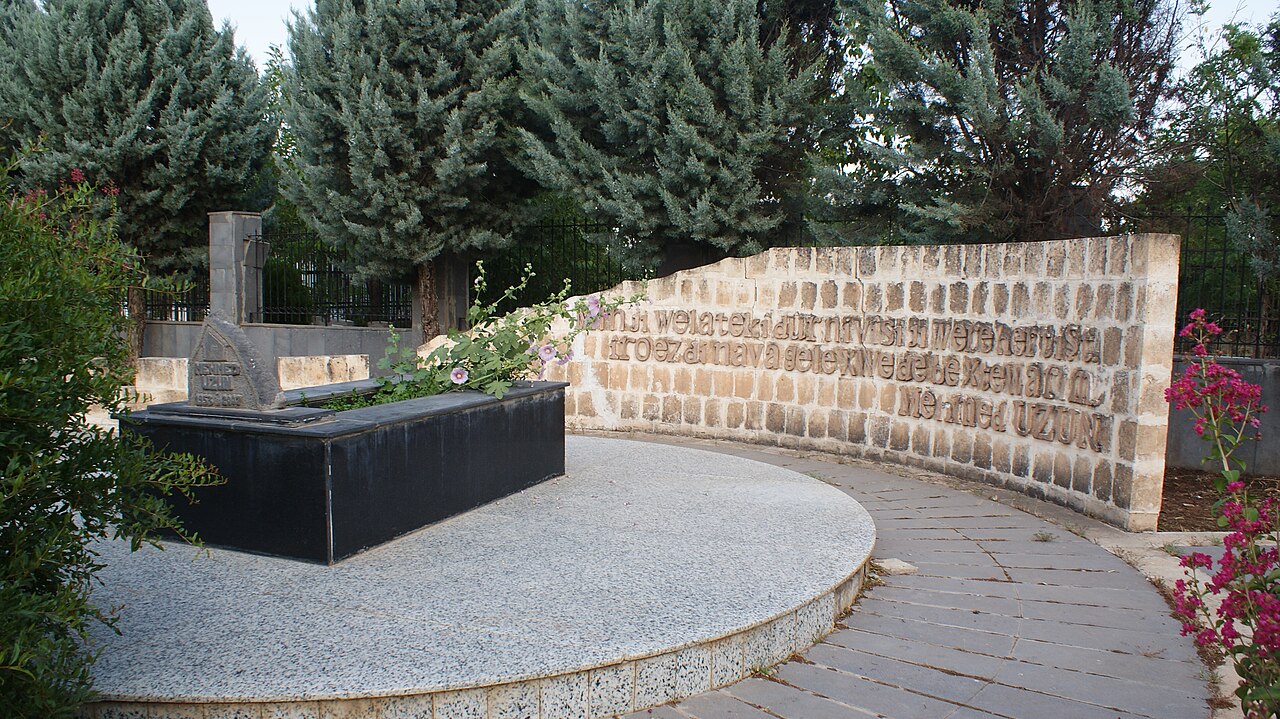 Tombe sombre d’Uzun devant un mur courbe portant une inscription kurde et des arbres