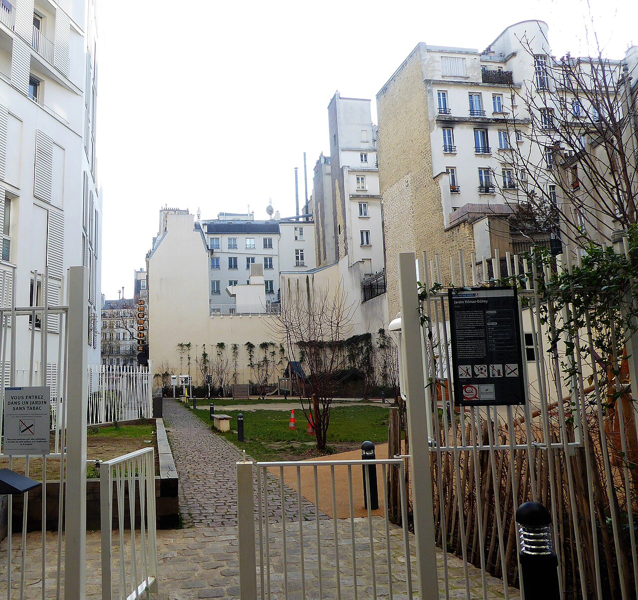 Paris’te Yılmaz Güney bahçesine açılan kapı ve taş döşeli yol; isim tabelası, çim ve apartmanlar arasında yapraksız ağaçlar